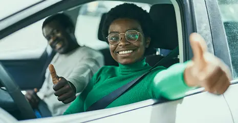 Mulher e homem sentados dentro de um carro sorrindo e olhando para fora enquanto fazem o sinal de positivo com suas mãos, com o polegar levantado, em sinal de aprovação