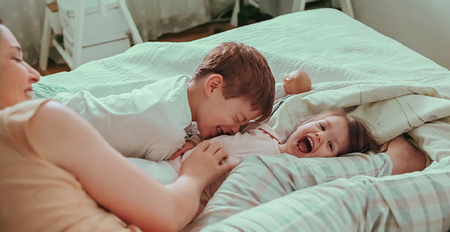 Foto de mãe fazendo cócegas em seus filhos na cama.