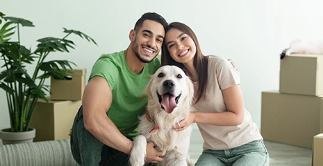 Homem e mulher abraçados junto à um cachorro de pelos claros sorrindo e olhando para a câmera. Estão sentandos em um sofá