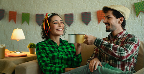 Homem e mulher com roupa xadrez sorrindo brindando com canecas em ambiente com bandeiras de Festa Juninas