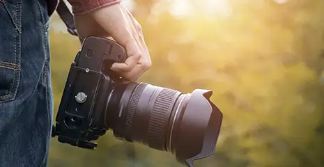 Foto de uma mão segurando uma câmera fotográfica profissional com lente grande acoplada no dispositivo. Ao fundo, pode-se ver árvores e a luz alaranjada do sol.