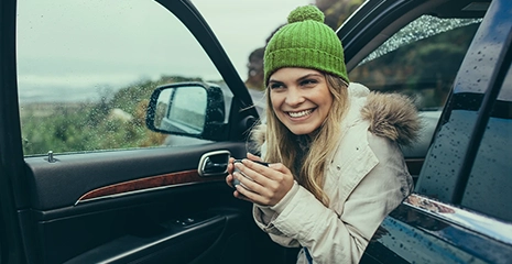 foto de mulher sentada no banco do motorista de uma carro, mas com as pernas para fora e a porta aberta. Ela está sorrindo e usando uma touca verde, enquanto segura uma bebida quente