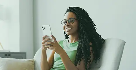 Mulher sorrindo enquanto mexe em celular sentada em um sofá, em ambiente interno