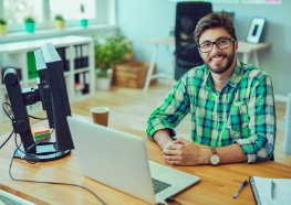 Homem com barba sorrindo, usando camisa xadrez verde e óculos, sentado em uma mesa de escritório com um MacBook